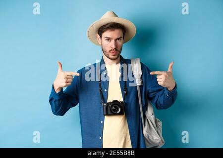 Giovane uomo sicuro e pronto che indica se stesso, auto-promozione o show-off, andare in vacanza estiva, indossare cappello di paglia, tenere zaino con Foto Stock