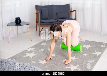 una giovane donna in un jumpsuit sportivo rosa in piedi le ginocchia si massaggiano le gambe con una schiuma verde rullo all'interno di una casa Foto Stock