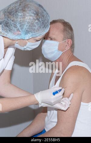 Vaccinazione contro il coronavirus. Un'infermiera dà il vaccino ad un uomo maturo che indossa una T-shirt bianca e uno scudo facciale. Foto Stock