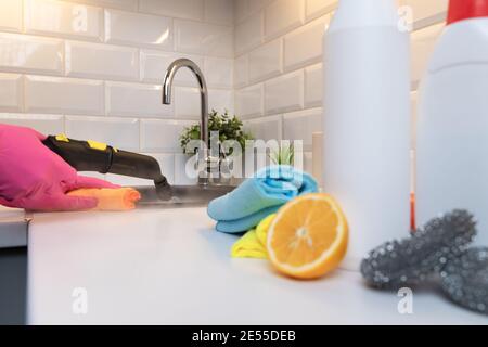 Casalinga in guanti di gomma pulizia lavello da cucina nero con un pulitore a vapore caldo e prodotti di pulizia sfocati, citron e spugne su piano cucina Foto Stock