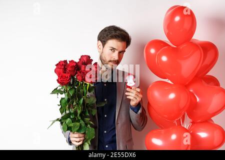 Bel ragazzo in tuta facendo proposta il giorno degli amanti, tenendo anello di impegno e rose rosse, preparare fiori a sorpresa e palloncini cuore per Foto Stock