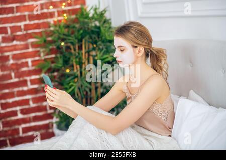 Bionda ragazza con una maschera sul viso seduta dentro letto con smartphone in mano Foto Stock