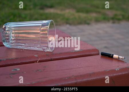 Un bicchiere svuotato e una sigaretta fumatrice gettata su un panca marrone e su uno sfondo di erba verde e. una passerella grigia Foto Stock