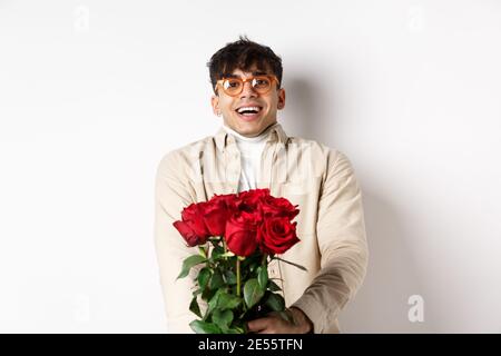 Uomo innamorato tenendo le rose rosse e guardando con tenerezza la macchina fotografica, fissando l'amante con il volto felice, celebrando San Valentino con la ragazza, in piedi Foto Stock