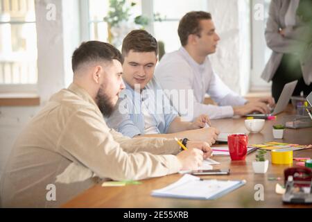 Discutere del progetto. Colleghi con vista laterale che lavorano insieme in un ufficio utilizzando dispositivi moderni durante le riunioni creative. Cancelleria, laptop, documenti. Concetto di business, ufficio, finanza, spazio aperto. Foto Stock