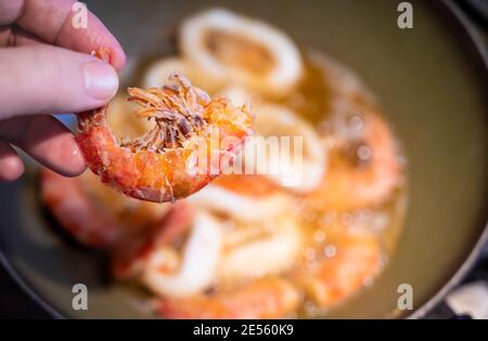 Home LIFE scene: Sullo sfondo gamberetti infarinati e anelli di calamari fritti in olio bollente. In primo piano, un gamberetto perfettamente cotto Foto Stock