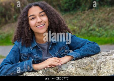 Ritratto esterno di bella ragazza afro-americana mista di razza felice femmina adolescente bambino sorridente con denti perfetti indossando giacca in denim appoggiandosi Foto Stock