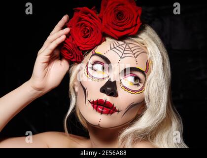 Splendido stile di Halloween Make-Up. Biondo modello indossare zucchero Skull Makeup con rose rosse, toni pelle pallido e capelli onde. Concetto di Santa Muerte Foto Stock