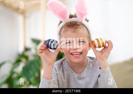 Felici i bambini di Pasqua. Ragazzo in orecchie conigliate di coniglio sulla testa con uova colorate a casa. Allegro pazzo sorridente bambino. Foto Stock