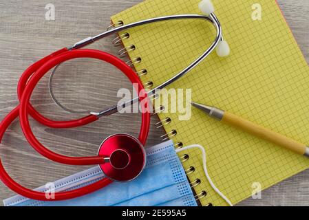 Tema medico ancora vita con uno stetoscopio rosso colorato e. maschera chirurgica posizionata sul lato del filo legato blocco note e penna visti come una posa piatta Foto Stock