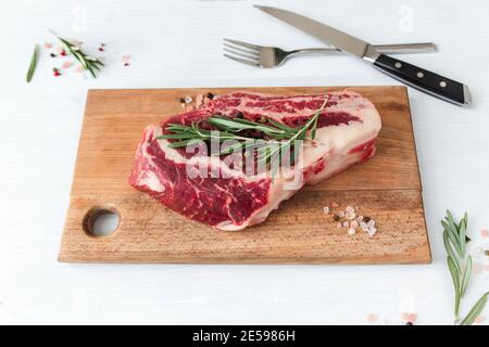 Bistecca di ribeye cruda a bordo, coltello, forchetta, pepe, rosmarino e sale rosa dell'Himalaya Foto Stock