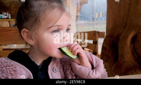 Carino bambina in caldo outerwear mordente fetta di fresco cetriolo mentre si siede al tavolo in un ristorante in stile rustico campagna Foto Stock