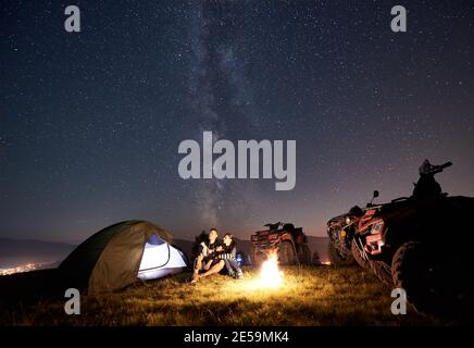 Felice coppia uomo e donna trevelers seduta vicino tenda turistica, fuoco ardente, tre moto quad atv sulla cima della montagna, godendo di una bella vista del cielo notturno pieno di stelle e Via Lattea Foto Stock