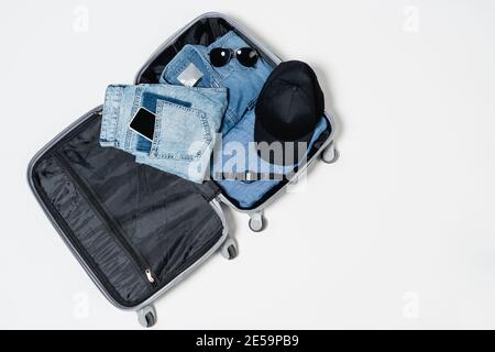vista dall'alto della valigia con capi in denim, cappuccio nero, smartphone, occhiali da sole e orologio da polso elettronico su bianco Foto Stock