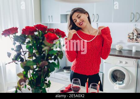 Giovane donna ha trovato bouquet di rose con candele, vino e dono in cucina. La ragazza felice prova sulla collana della perla. San Valentino sorpresa Foto Stock