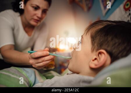 Un bambino malato che si trova a letto e prende un medicinale dalla madre. Una madre che offre la medicina su un cucchiaio al suo figlio malato. Foto Stock