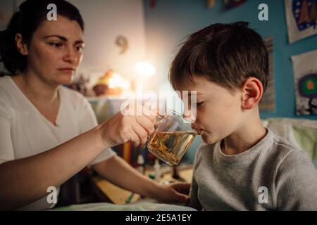 Un bambino si sente malbene a bere un tè alle erbe offerto dalla madre. Un ritratto di un ragazzo malato seduto nel suo letto di notte sorseggiando una bevanda calda. Foto Stock
