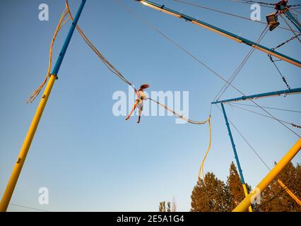 Una bambina allegra vola su fasce elastiche luminose e elastiche e salta su un trampolino godendo la vacanza attesa a lungo in il sole caldo Foto Stock