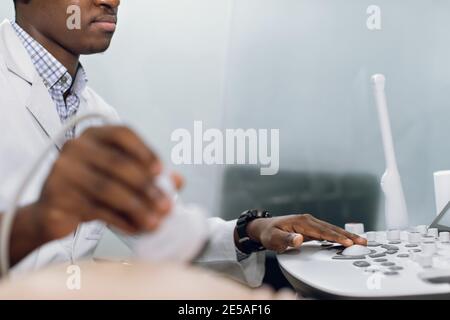 Primo piano inquadratura delle mani di un medico africano di sesso maschile, screening della pancia incinta con trasduttore a ultrasuoni, pulsanti sul pannello di controllo. Messa a fuoco Foto Stock