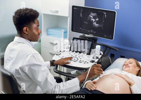 Felice giovane donna caucasica incinta che ha ecografia diagnostica in clinica moderna, guardando il suo bel nero uomo medico ostetrista, mettendo Foto Stock