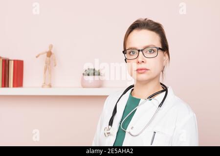 Ritratto di sicuro giovane dottore con giacca bianca con stetoscopio e auricolare in piedi nel suo ufficio domestico guardando telecamera pronta per onl Foto Stock