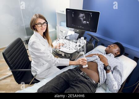 Giovane donna medico ecografo che utilizza ecografo al lavoro, tenendo in mano scanner ecografico mentre esamina l'addome di un paziente africano maschio a. Foto Stock