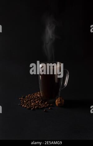 Caffè nero caldo in una tazza di vetro con tartufo al cioccolato e molti chicchi di caffè su sfondo scuro. Concetto di amore per il caffè per la colazione. Vista frontale Foto Stock