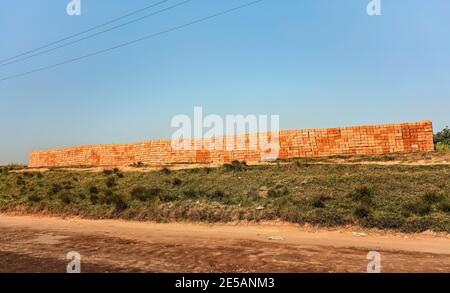 Mattoni rossi arancioni fatti di argilla locale disposti in un ampio stack, pronti per costruire una casa vicino alla strada Foto Stock