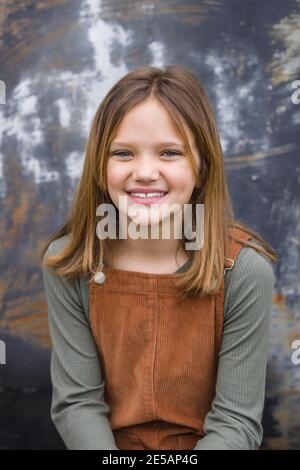 Giovane ragazza in un vestito marrone generale in inverno seduto vicino a un muro di un magazzino urbano Foto Stock