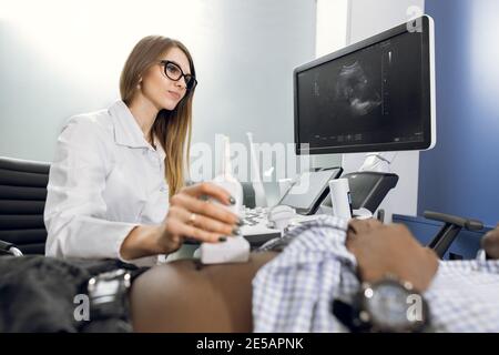 Il medico femminile focalizzato esegue l'esame ecografico degli organi interni di il suo paziente maschio nero per la diagnosi di disagio dello stomaco a. il moderno Foto Stock