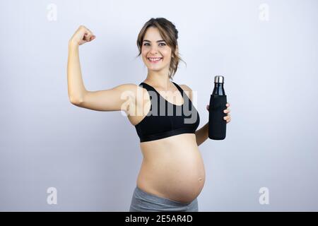 Giovane sportivo incinta con indosso sportivo che tiene una bottiglia con acqua sfondo bianco che mostra i muscoli delle braccia sorridendo orgoglioso Foto Stock