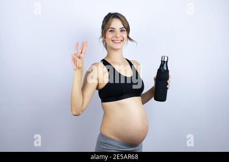 Giovane sportivo incinta con indosso sportivo che tiene una bottiglia con acqua sfondo bianco che mostra e punta verso l'alto con le dita numero tre mentre Foto Stock