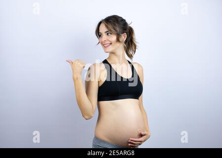 Giovane sportivo incinta indossando abbigliamento sportivo su sfondo bianco sorridente e. puntare con la mano e il dito verso il lato Foto Stock