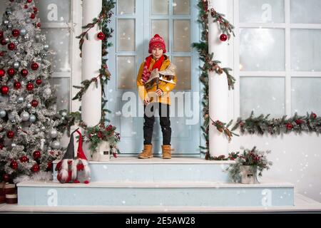 Bel ragazzino a natale. Il bambino divertente tiene vecchi pattini e si leva sul portico di una casa decorata con le decorazioni dell'albero di Natale Foto Stock