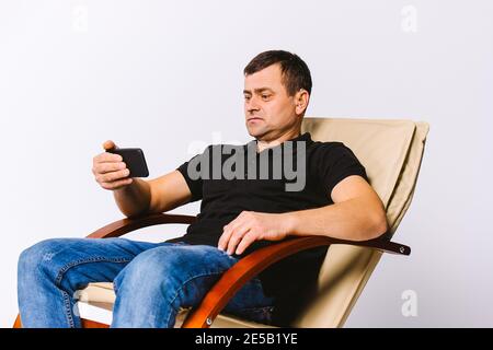 Uomo anziano non udente con un apparecchio acustico seduto in una poltrona in pelle che guarda un film al telefono. Sfondo bianco. Foto Stock