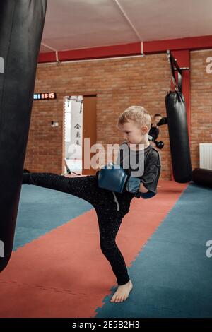 Ritratto di un ragazzo che calcia e pugni un sacchetto di boxe in palestra. Bambino in un sportswear che indossa i guanti di boxing che migliorano le sue abilità di kickboxing. Foto Stock