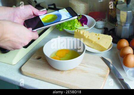 Una donna scatta foto di uova in una ciotola sul suo telefono nella sua cucina. Cibo blogger. Concetto di social media. Orientamento orizzontale. Foto Stock