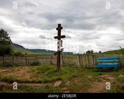 Una vecchia croce di legno si erge lateralmente vicino ad una recinzione storta e ad una panchina sullo sfondo di un villaggio in una giornata estiva nuvolosa. Foto Stock