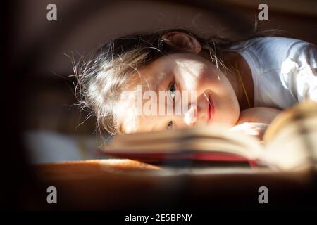 primo piano ritratto attraverso le barre di un letto in ghisa di una ragazza sorridente sdraiata faccia in giù su un libro aperto alla luce del sole della camera da letto Foto Stock