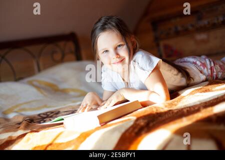 primo piano una ragazza sorridente e felice si trova sul suo stomaco su una coperta sul letto e legge un libro nella camera da letto di una cabina di legno nel villaggio Foto Stock