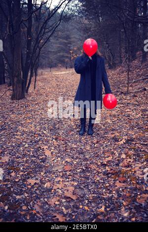 donna in camice nero in piedi nella foresta in tenuta di autunno un palloncino rosso sul viso Foto Stock