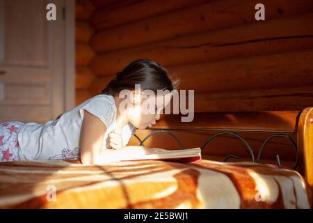 una ragazza di cinque anni si trova sullo stomaco su una coperta sul letto e legge un libro in camera da letto di una casa di villaggio di tronchi alla luce del sole Foto Stock