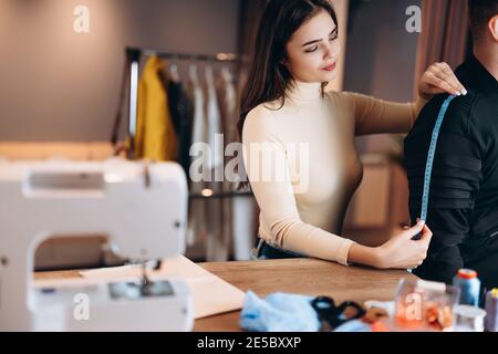 Giovane cameriera con macchina da cucire. Misure di sforzo di mare per l'uomo in officina. Creazione di corsi online di design di abbigliamento. Foto Stock