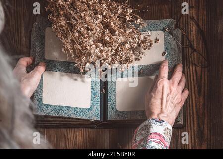 Mani rughe di una donna anziana dai capelli grigi che tiene un album fotografico d'epoca, accanto al bouquet di fiori secchi. Vista dall'alto. Concetto di nostalgia, ricordi Foto Stock