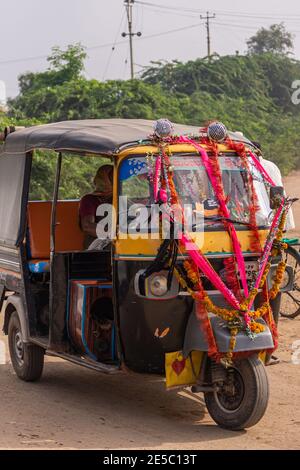 Nandakeshwar, Karnataka, India - 7 novembre 2013: Closeup di taxi triciclo giallo-nero su strada sterrata con una paasenger femminile sotto il cielo argentato. Verde fo Foto Stock