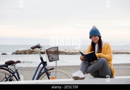 Donna seduta di fronte al mare che legge un libro e in bicicletta Foto Stock