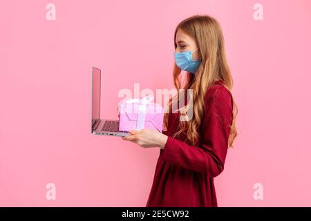 Felice elegante giovane donna in abito rosso, indossa una maschera medica, utilizza un computer portatile, contiene un regalo per San Valentino, su uno sfondo rosa, San Valentino c Foto Stock