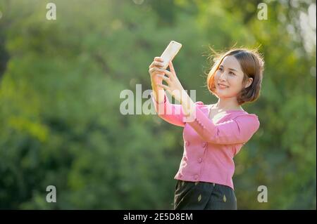 Donna sorridente in piedi in un giardino prendendo un selfie con il suo telefono cellulare, Thailandia Foto Stock