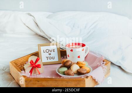 Colazione di San Valentino a letto per amante. Carte d'amore nella cornice, una tazza di caffè o cacao, macaroon, scatola regalo a forma di cuore con nastro rosso sopra Foto Stock