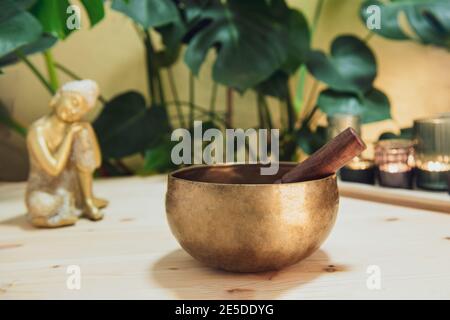 Ciotola tibetana con bastone speciale, candele brucianti e statuetta decorativa del Buddha dorato sul tavolo di legno con piante verdi di fondo Foto Stock